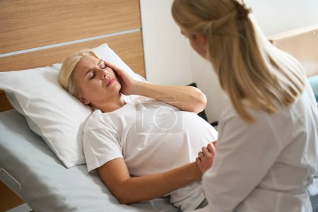 Téléchargez les photos : Infirmière tenant la main d'une femme enceinte caucasienne adulte avec douleur à la tête sur un lit médical à la maternité - en image libre de droit