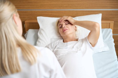 Foto de Enfermera parcial mirando a mujer embarazada caucásica adulta con dolor de cabeza en cama médica en el hospital de maternidad - Imagen libre de derechos