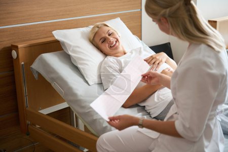 Foto de Sonriente mujer embarazada caucásica adulta acostada en la cama médica y mirando a la enfermera revisando su electrocardiograma en el hospital de maternidad - Imagen libre de derechos