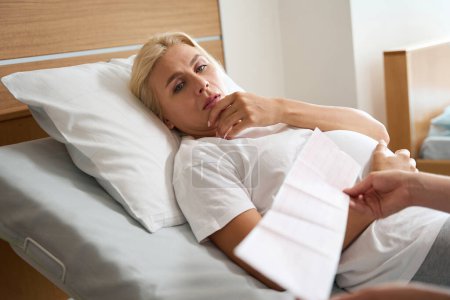 Foto de Mujer embarazada caucásica adulta grave acostada en la cama médica y mirando su electrocardiograma en manos de una enfermera parcial en el hospital de maternidad - Imagen libre de derechos