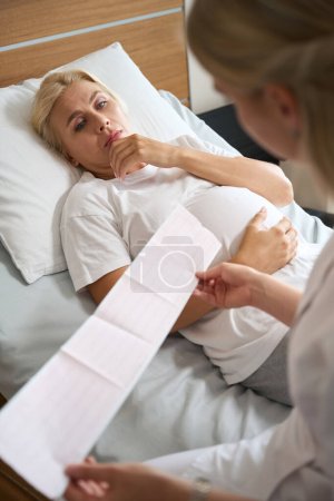 Téléchargez les photos : Femme enceinte caucasienne adulte allongée sur un lit médical et regardant son électrocardiogramme dans les mains d'une infirmière à la maternité - en image libre de droit
