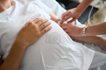 Téléchargez les photos : Concentration sélective du ventre de la femme enceinte cultivée tandis que l'infirmière partielle floue mettre compteur de gouttes à elle à la maternité - en image libre de droit