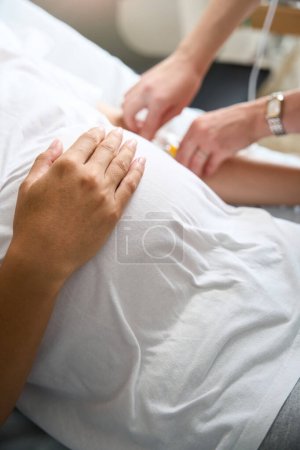 Téléchargez les photos : Image partielle de l'infirmière floue mettre comptoir de gouttes à la femme enceinte sur le lit médical dans la maternité - en image libre de droit