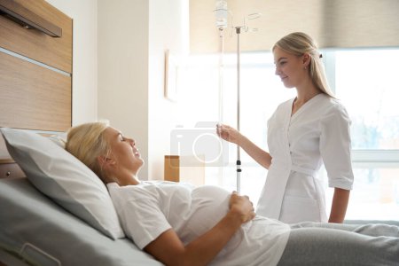 Photo for Side view of smiling european female nurse put drop counter to adult caucasian pregnant woman with closed eyes on medical bed in maternity hospital - Royalty Free Image