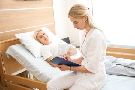 Foto de Sonriente mujer embarazada caucásica adulta acostada en la cama médica y mirando a la enfermera europea escribiendo en el portapapeles mientras la revisa en el hospital de maternidad - Imagen libre de derechos