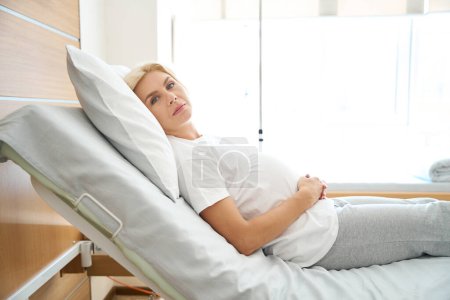 Foto de Vista lateral de una mujer embarazada europea adulta grave acostada en la cama médica, abrazando su vientre y mirando a la cámara en el hospital de maternidad - Imagen libre de derechos
