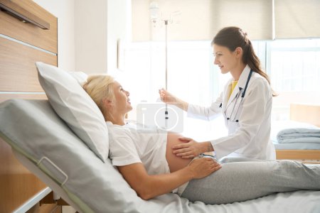 Foto de Médico caucásico femenino serio que comprueba el vientre embarazada de la mujer europea en la cama médica y pone el contador de la gota a ella en la clínica de maternidad - Imagen libre de derechos