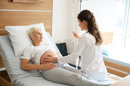 Foto de Mujer europea mirando al médico caucásico mujer revisando su vientre embarazada y poner contador de gota en la clínica de maternidad - Imagen libre de derechos