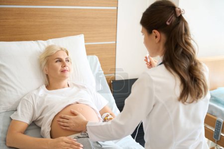 Foto de Mujer europea y mujer caucásica médico control de vientre embarazada y poner contador de gota a la paciente y se miran entre sí en la clínica de maternidad - Imagen libre de derechos