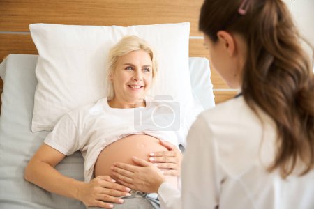 Foto de Mujer europea sonriente mirando a una enfermera revisando su vientre embarazada en la clínica de maternidad - Imagen libre de derechos