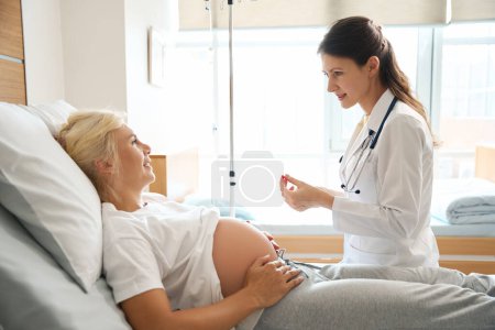Téléchargez les photos : Infirmière européenne préparant la seringue pour injection et femme caucasienne enceinte sur le lit médical se regardant dans la maternité - en image libre de droit