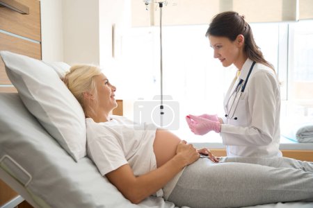 Téléchargez les photos : Infirmière européenne préparant la seringue pour injection et souriante femme caucasienne enceinte sur le lit médical se regardant dans la maternité - en image libre de droit