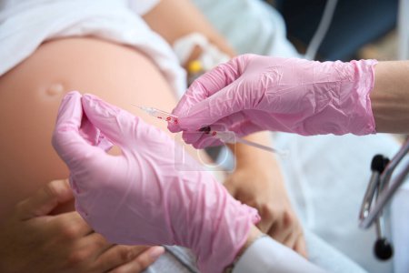 Foto de Imagen parcial de las manos enfermeras en guantes de látex que preparan la jeringa para inyección en el vientre embarazada de una paciente femenina borrosa en la clínica de maternidad - Imagen libre de derechos