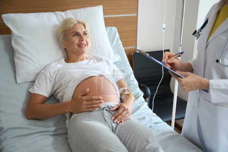 Téléchargez les photos : Cropped femme médecin écrit dans le presse-papiers sur souriant adulte caucasien femme enceinte sur lit médical tout en vérifiant son dans la maternité - en image libre de droit