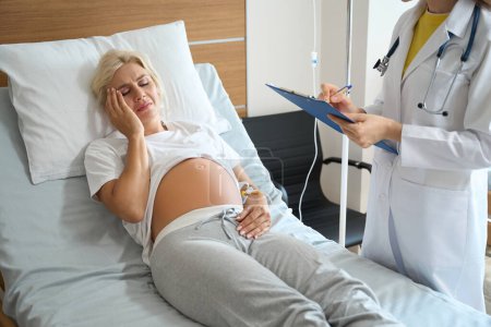 Foto de Doctora parcial escribiendo en portapapeles sobre una mujer embarazada caucásica adulta con dolor de cabeza en la cama médica mientras la revisa en el hospital de maternidad - Imagen libre de derechos