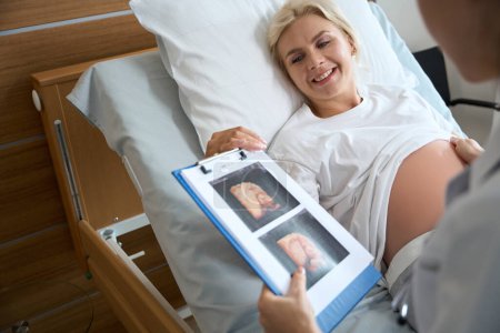 Téléchargez les photos : Souriant adulte caucasien femme enceinte sur le lit médical en regardant la photo échographie de son futur bébé en main de la femme cultivée médecin à la maternité - en image libre de droit