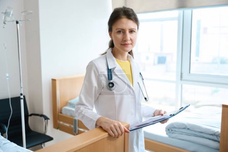 Foto de Vista frontal de una doctora caucásica seria que usa un abrigo blanco con portapapeles mirando a la cámara en la clínica - Imagen libre de derechos