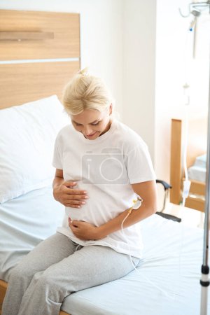 Photo for Smiling adult european pregnant woman sitting on medical bed and embracing her belly in maternity hospital - Royalty Free Image