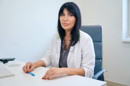Foto de Médico europeo adulto serio vistiendo bata blanca sentado en el escritorio y mirando a la cámara en la clínica - Imagen libre de derechos