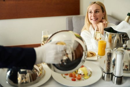 Téléchargez les photos : Serveur en gants blancs sert le petit déjeuner à un invité de l'hôtel, une femme apprécie une coupe de champagne - en image libre de droit