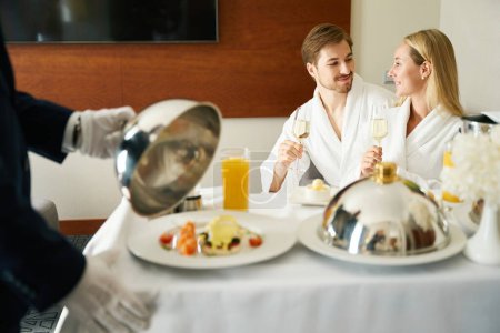 Téléchargez les photos : Serveur servi le petit déjeuner aux jeunes mariés dans leur chambre sur une table mobile, un homme et une femme profiter du champagne - en image libre de droit