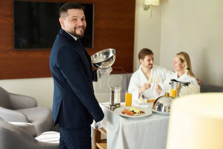 Téléchargez les photos : Serveur souriant servi le petit déjeuner aux jeunes mariés dans leur chambre, un homme et une femme dégustant du champagne - en image libre de droit