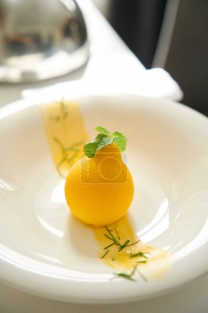 Téléchargez les photos : Dessert jaune est servi avec des feuilles de menthe sur une assiette blanche, couvercle cloche se trouve à côté sur la table de service - en image libre de droit