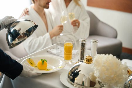 Téléchargez les photos : Employé de l'hôtel servi le petit déjeuner aux jeunes mariés dans leur chambre, un homme et une femme profiter du champagne - en image libre de droit