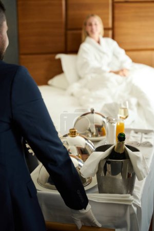 Foto de El camarero sirve el desayuno en la habitación con champán en la mesa de servir, un huésped del hotel con una acogedora bata de felpa. - Imagen libre de derechos