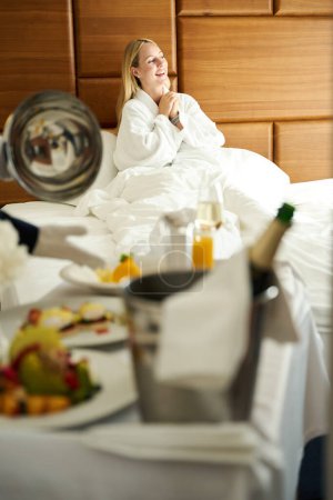 Téléchargez les photos : Serveur sert le petit déjeuner à une invitée de l'hôtel dans sa chambre, femelle dans un peignoir éponge confortable - en image libre de droit
