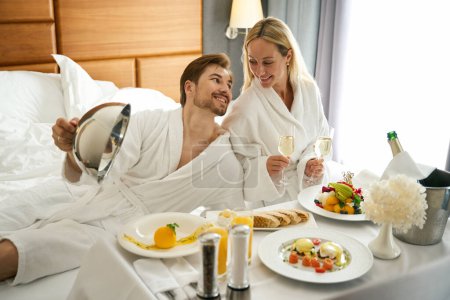 Téléchargez les photos : Guy et sa copine ont commandé le petit déjeuner avec du champagne dans leur chambre, ils se sont installés sur un grand lit - en image libre de droit