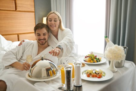 Téléchargez les photos : Homme et femme dégustent un petit déjeuner au champagne assis sur un grand lit - en image libre de droit