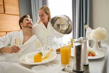 Photo for Happy newlyweds enjoy a champagne breakfast, they are seated on a large bed - Royalty Free Image