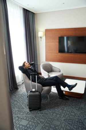 Foto de El hombre sonriente se sienta con los brazos extendidos en una habitación de hotel, junto a una maleta de viaje - Imagen libre de derechos