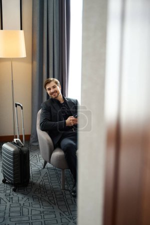 Photo for Smiling male sitting with a mobile phone in a hotel room, next to a travel suitcase - Royalty Free Image