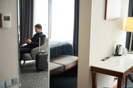 Téléchargez les photos : Homme en costume de voyage assis avec un téléphone portable dans la chaise, à côté d'une valise de voyage - en image libre de droit