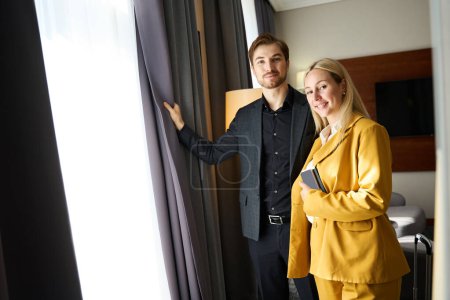Photo for Traveling married couple standing by the window in a hotel room, woman in a yellow travel suit - Royalty Free Image