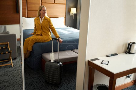 Foto de Mujer con un traje de viaje amarillo se sienta en una cama grande en una habitación de hotel, junto a su maleta de viaje - Imagen libre de derechos