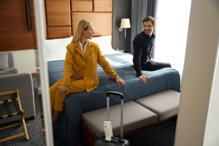 Photo for Traveling couple relaxing on bed in hotel room, woman in yellow travel suit - Royalty Free Image