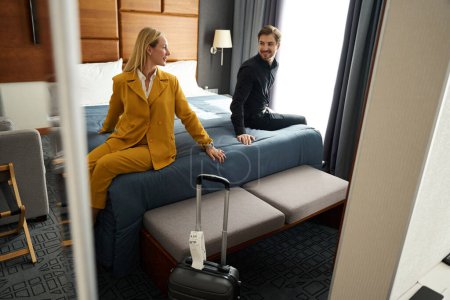 Téléchargez les photos : Voyager couple marié se détendre sur le lit dans la chambre d'hôtel, femme en costume de voyage jaune - en image libre de droit