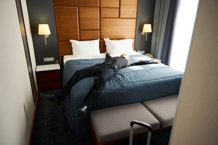 Téléchargez les photos : Heureux jeune homme couché les bras tendus sur une couverture bleue, la chambre est propre et confortable - en image libre de droit