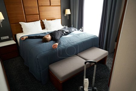 Téléchargez les photos : L'homme se couche les bras tendus sur un grand lit dans une chambre d'hôtel, il porte des vêtements de voyage noirs - en image libre de droit