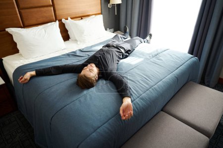 Téléchargez les photos : Le gars se couche les bras tendus sur un grand lit dans une chambre d'hôtel, il est en tenue décontractée noire - en image libre de droit