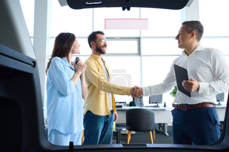 Photo for Manager greets clients at a car dealership, people stand by a car with an open trunk - Royalty Free Image