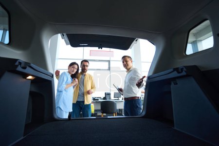 Foto de Gerente consulta a los clientes en un concesionario de automóviles, que están al lado de un coche con un baúl abierto - Imagen libre de derechos