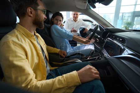 Foto de Gerente en un concesionario de automóviles presenta un coche nuevo a los clientes, una pareja se sienta en el interior de los coches - Imagen libre de derechos
