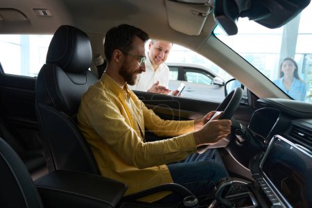 Hombre satisfecho en un coche está estudiando la funcionalidad de un modelo popular, con un gerente-consultor cercano