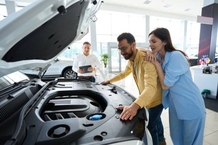 Photo for Man and a woman are inspecting a car under the hood, they are being advised by a customer service manager - Royalty Free Image