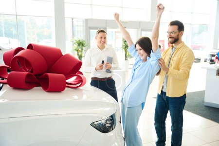 Woman rejoices at car given to her with a red bow on the hood, next to her husband and consultant