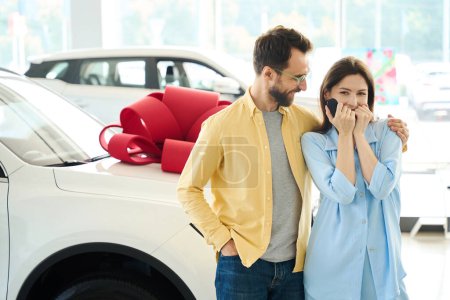 Happy woman rejoices at a gifted car, her husband hugs her shoulders